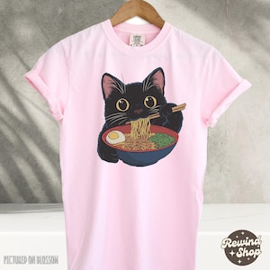 May include: Pink t-shirt with a black cat eating ramen noodles. The cat is wearing a red collar and has a bowl of ramen in front of it. The text "Comfy Clothes" is on the tag.