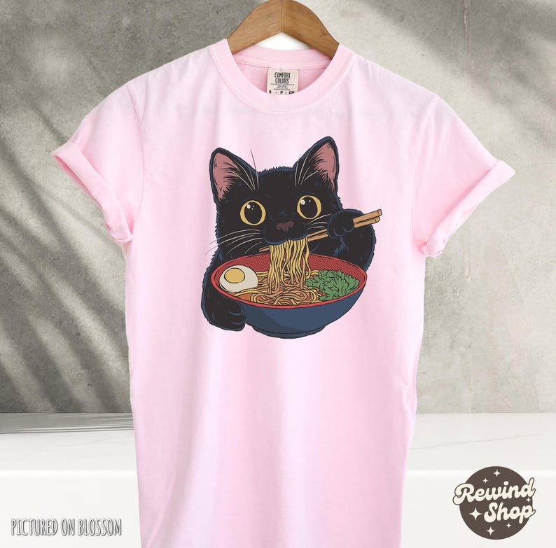 May include: Pink t-shirt with a black cat eating ramen noodles. The cat is wearing a red collar and has a bowl of ramen in front of it. The text "Comfy Clothes" is on the tag.