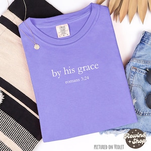 May include: A lilac Comfort Colors t-shirt with the text "by his grace romans 3:24" printed in white. The shirt is laid flat with a gold necklace, denim shorts, and a black and white striped textile.