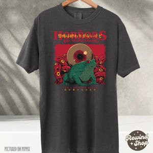 Puede incluir: Una camiseta gris oscuro con un estampado gráfico de una rana verde con un ojo grande y tentáculos. El texto "The Horrors Are Never Ending" está impreso en rojo sobre la rana. El texto japonés "恐怖は終わりがないけど" está impreso en rojo sobre el texto inglés.