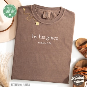 May include: A brown t-shirt with the text "by his grace romans 3:24" in white. A gold necklace with a pendant is placed on the shirt. A beige hat and brown sandals are also visible.