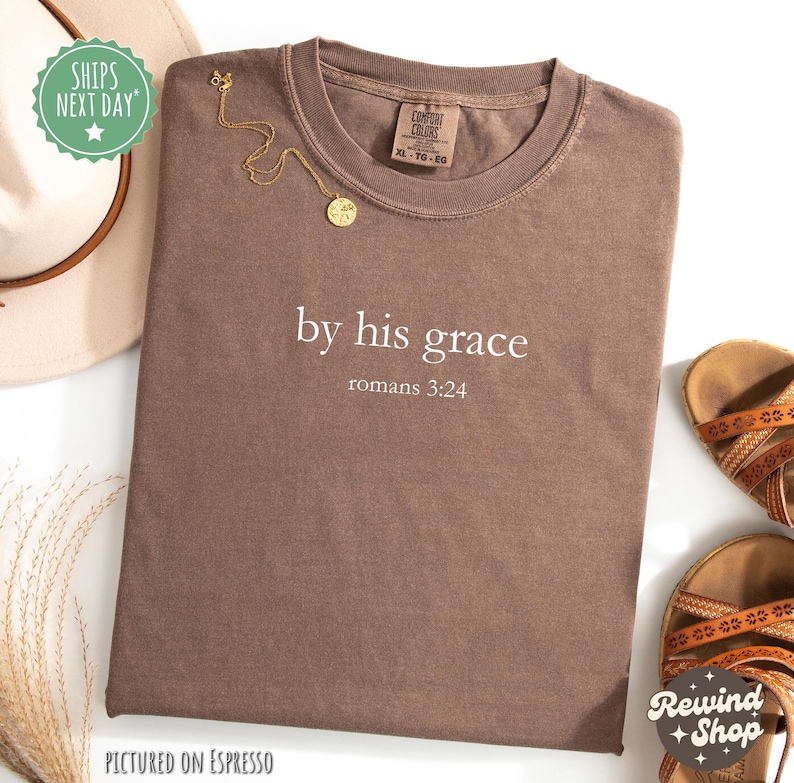 May include: A brown t-shirt with the text "by his grace romans 3:24" in white. A gold necklace with a pendant is placed on the shirt. A beige hat and brown sandals are also visible.