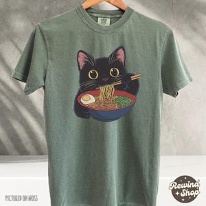 May include: A green t-shirt with a black cat eating ramen noodles. The cat has big, round eyes and is holding chopsticks. The text "Rewind Shop" is in the bottom right corner of the image.