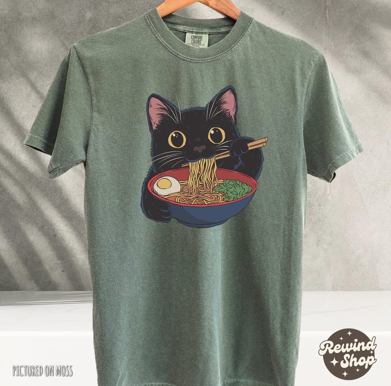 May include: A green t-shirt with a black cat eating ramen noodles. The cat has big, round eyes and is holding chopsticks. The text "Rewind Shop" is in the bottom right corner of the image.