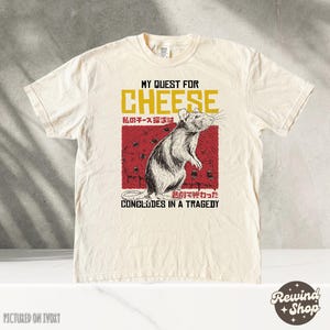 Op de afbeelding: Een beige t-shirt met een afbeelding van een rat en kaas. De tekst op het shirt luidt "My Quest for Cheese" en "My cheese quest concludes in a tragedy".