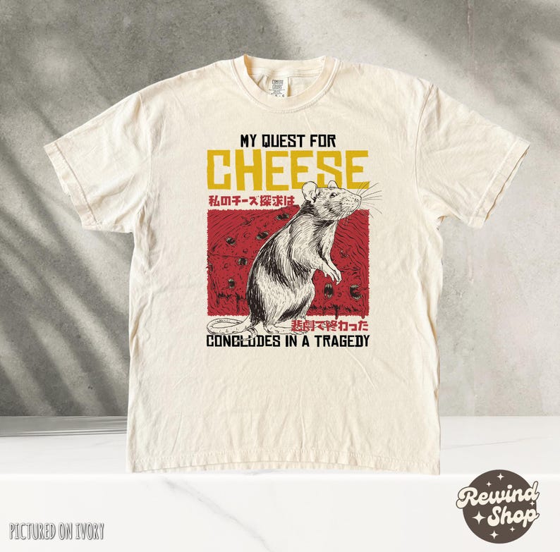 Op de afbeelding: Een beige t-shirt met een afbeelding van een rat en kaas. De tekst op het shirt luidt "My Quest for Cheese" en "My cheese quest concludes in a tragedy".