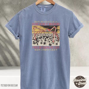 May include: A blue t-shirt with rolled-up sleeves. The shirt features a graphic of a spider-like creature with multiple eyes and text in English and Japanese. The shirt is displayed on a hanger.