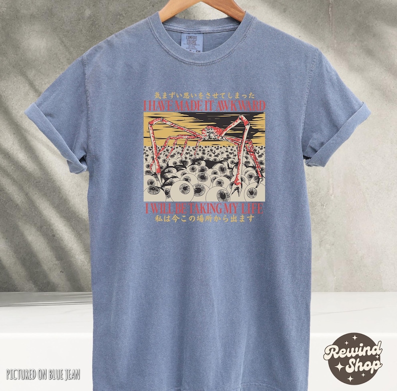May include: A blue t-shirt with rolled-up sleeves. The shirt features a graphic of a spider-like creature with multiple eyes and text in English and Japanese. The shirt is displayed on a hanger.