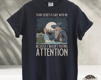 Op de afbeelding: Een donkerblauw T-shirt met een afbeelding van een tovenaar met een lange witte baard en een puntige hoed. De tovenaar staat in een bos met een kikker op een boomstam. De tekst op het shirt luidt "Je geheim is veilig bij mij, omdat ik niet opletten."
