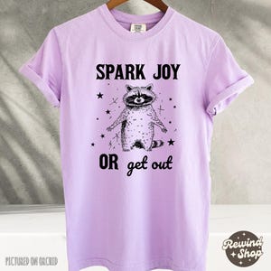 以下が含まれることがあります： ラベンダー色のTシャツに、黒と白のラクーンドローイングと「Spark Joy or get out」の文字がプリントされています。
