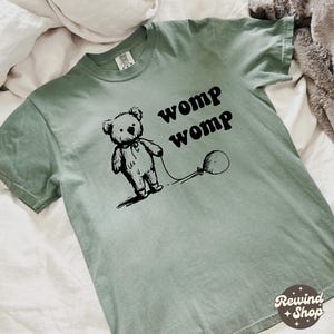Puede incluir: Camiseta verde salvia con una ilustración negra de un oso de peluche sosteniendo un globo y las palabras "womp womp". La camiseta tiene cuello redondo y mangas cortas. El logo de Rewind Shop está en la esquina inferior derecha.