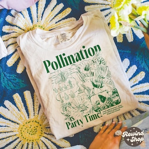 May include: A cream-coloured t-shirt featuring the word "Pollination" in green, a botanical illustration, and the phrase "Party Time!" The shirt also includes the text "Just add flowers, bees, and a little shake-shake from the wind!"