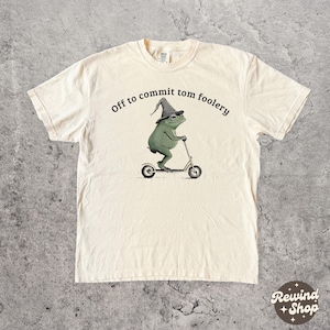 Può includere: T-shirt color crema con un ranocchio dei cartoni animati su uno scooter, con un cappello da mago e occhiali. Il testo "Off to commit tom foolery" è arcuato sopra il ranocchio. Il logo Rewind + Shop è in basso a destra.