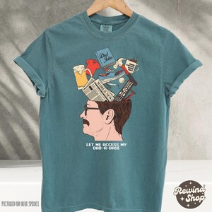 May include: Teal blue t-shirt with a graphic of a man's head with various items emerging from it, including a beer, a remote, and a newspaper. The text "LET ME ACCESS MY DAD-A-BASE" is below the illustration. The shirt is hanging on a wooden hanger.