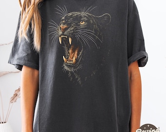 May include: Dark gray t-shirt featuring a detailed illustration of a roaring black panther. The panther's open mouth reveals sharp teeth and a tan interior. The shirt has short sleeves and a relaxed fit. The text "Rewind + Shop" is visible on the bottom right.