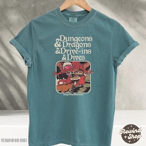 Może przedstawiać: Turkusowa koszulka z podwiniętymi rękawami. Koszulka zawiera napis "Dungeons & Dragons & Drive-ins & Dives" nad grafiką przedstawiającą czerwonego smoka i osobę. Grafika jest oznaczona jako "Escape From Flavortown."