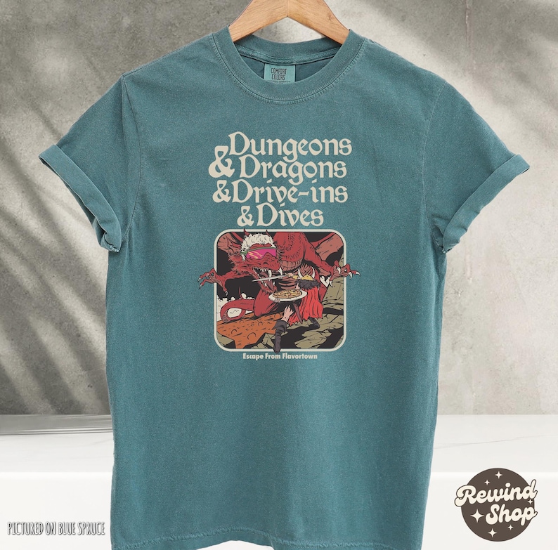 Może przedstawiać: Turkusowa koszulka z podwiniętymi rękawami. Koszulka zawiera napis "Dungeons & Dragons & Drive-ins & Dives" nad grafiką przedstawiającą czerwonego smoka i osobę. Grafika jest oznaczona jako "Escape From Flavortown."