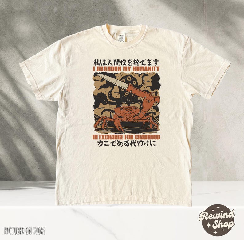 May include: Ivory-colored t-shirt with a graphic design featuring a crab holding a knife, with the text "I Abandon My Humanity" and "In Exchange For Crabhood". The shirt has a crew neck and short sleeves.