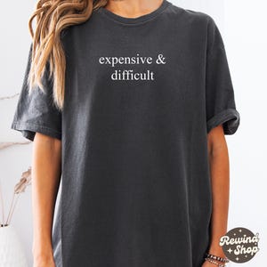 Può includere: Una maglietta grigia scura con la scritta "expensive & difficult" stampata in bianco sul davanti.