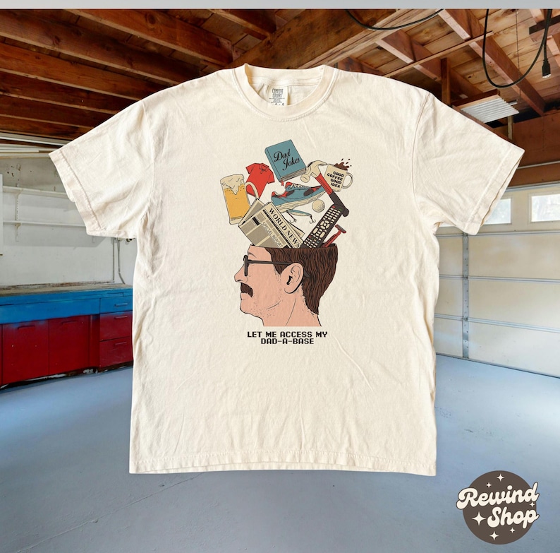 May include: Cream-colored t-shirt featuring a graphic of a man's head with various items emerging from it, including a newspaper, beer, and a remote control. The text "LET ME ACCESS MY DAD-A-BASE" is printed below the image.
