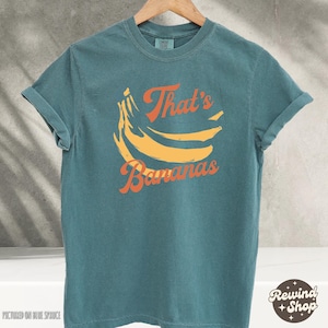 May include: Teal-colored t-shirt with rolled-up sleeves. The shirt features a graphic of yellow bananas and the text "That's Bananas" in orange. The shirt is hanging on a wooden hanger.