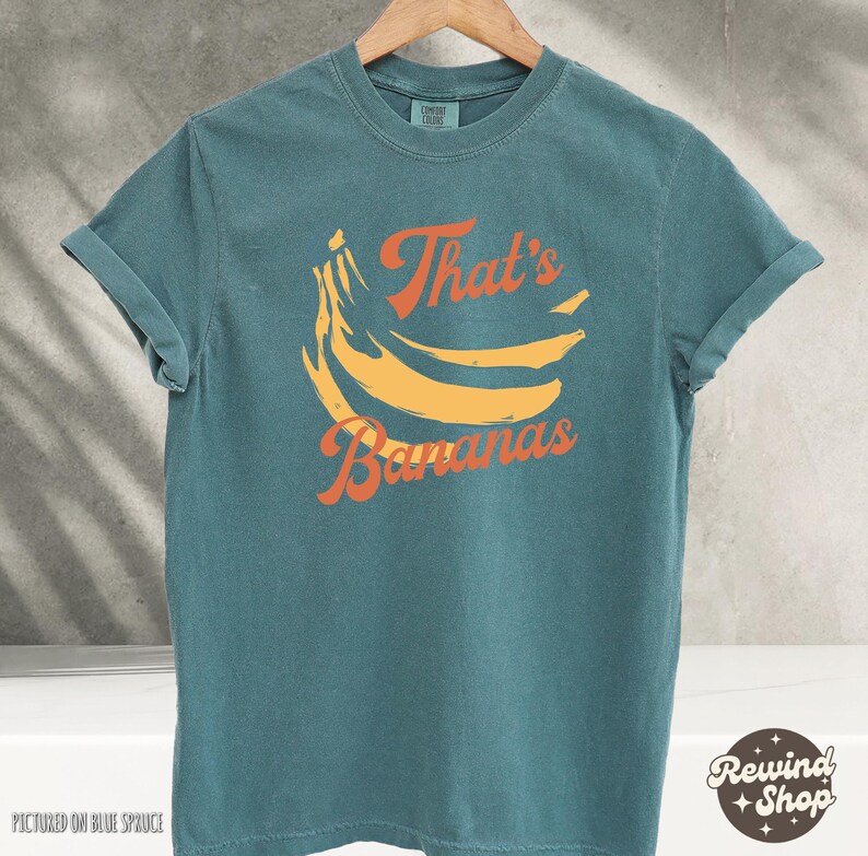 May include: Teal-colored t-shirt with rolled-up sleeves. The shirt features a graphic of yellow bananas and the text "That's Bananas" in orange. The shirt is hanging on a wooden hanger.