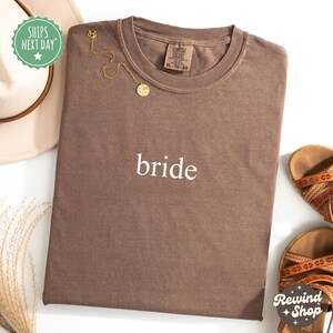 Pode incluir: Uma camiseta marrom com a palavra "bride" impressa em branco na frente. A camiseta é feita de um tecido macio e confortável e tem um caimento relaxado.