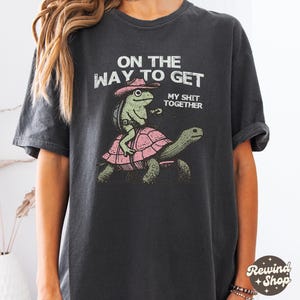 May include: A black t-shirt with a graphic of a frog wearing a cowboy hat riding a turtle. The text on the shirt reads "On the way to get my shit together."
