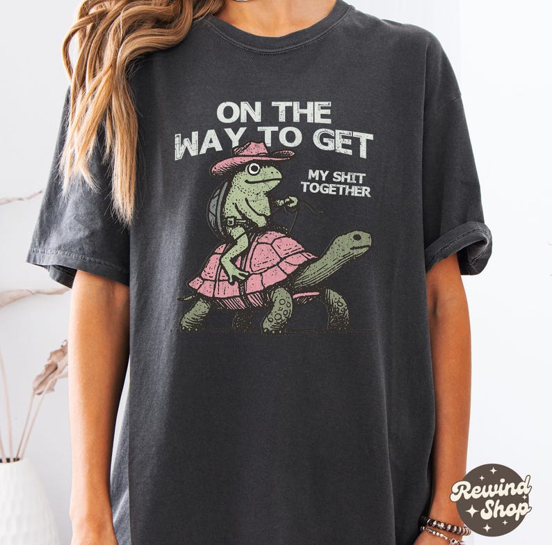 May include: A black t-shirt with a graphic of a frog wearing a cowboy hat riding a turtle. The text on the shirt reads "On the way to get my shit together."