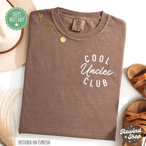 Könnte beinhalten: Ein braunes T-Shirt mit dem Aufdruck "COOL Uncles CLUB" in weiß. Das Shirt ist auf einem hellbraunen Hintergrund abgebildet.