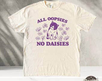Puede incluir: Una camiseta de color crema con un gráfico morado de un gato sentado con el texto "All oopsies no daisies" impreso en ella.