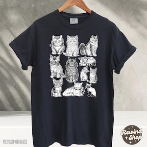 K&ouml;nnte beinhalten: Schwarzes T-Shirt mit einer wei&szlig;en Illustration verschiedener Katzenrassen in verschiedenen Posen. Das T-Shirt hat kurze &Auml;rmel und einen Rundhalsausschnitt. Der Text "Pictured on Black" befindet sich unten links.