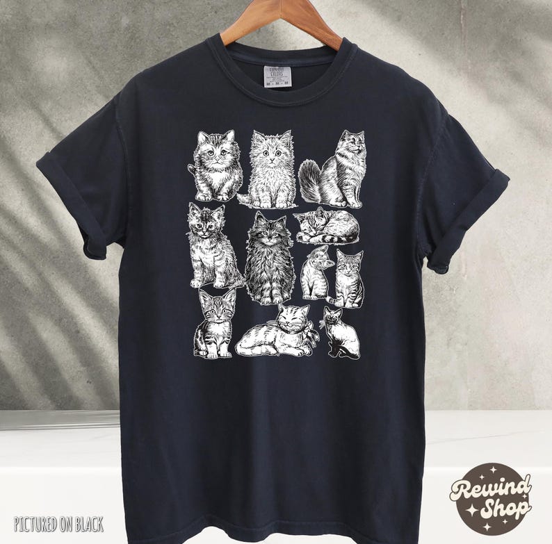 K&ouml;nnte beinhalten: Schwarzes T-Shirt mit einer wei&szlig;en Illustration verschiedener Katzenrassen in verschiedenen Posen. Das T-Shirt hat kurze &Auml;rmel und einen Rundhalsausschnitt. Der Text "Pictured on Black" befindet sich unten links.