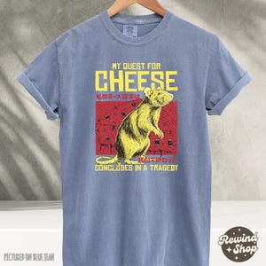 Op de afbeelding: Een blauw t-shirt met een afbeelding van een rat en de tekst "My Quest for Cheese" en "私のチーズ探求は...で終わっ" en "Concludes in a Tragedy".