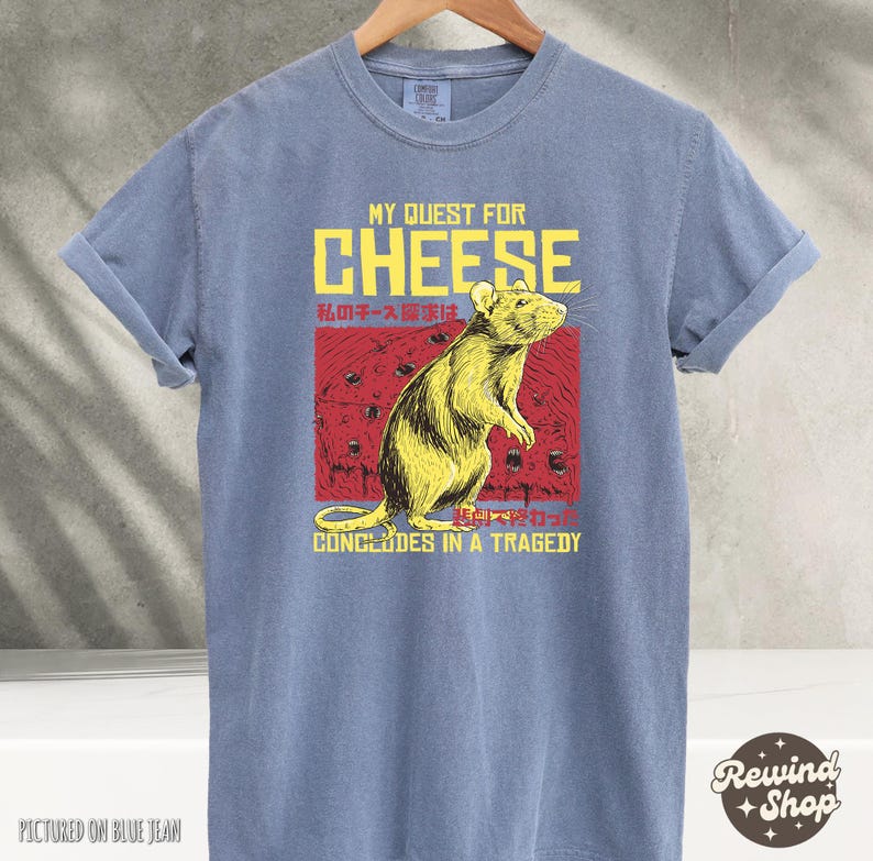 Op de afbeelding: Een blauw t-shirt met een afbeelding van een rat en de tekst "My Quest for Cheese" en "私のチーズ探求は...で終わっ" en "Concludes in a Tragedy".