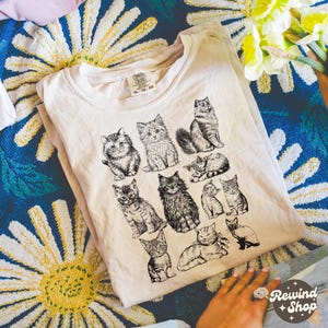 K&ouml;nnte beinhalten: Cremefarbenes T-Shirt mit einer Sammlung detaillierter Schwarz-Wei&szlig;-Katzenillustrationen. Das Shirt hat einen Rundhalsausschnitt und kurze &Auml;rmel. Das Etikett lautet "Comfort Colors" und "XL-TG-EG".