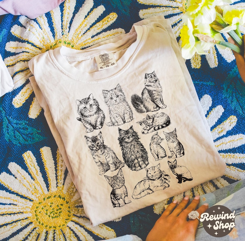 K&ouml;nnte beinhalten: Cremefarbenes T-Shirt mit einer Sammlung detaillierter Schwarz-Wei&szlig;-Katzenillustrationen. Das Shirt hat einen Rundhalsausschnitt und kurze &Auml;rmel. Das Etikett lautet "Comfort Colors" und "XL-TG-EG".