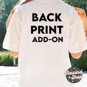 以下が含まれることがあります： クリーム色のTシャツ。太字の黒い文字で「BACK PRINT ADD-ON」と書かれています。Rewind + Shopのロゴが右下に表示されています。カジュアルなスタイルのシャツです。