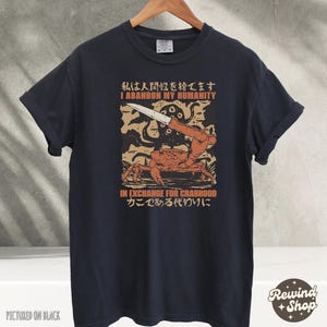 May include: Black t-shirt with a graphic featuring a crab holding a knife, with the text "I ABANDON MY HUMANITY IN EXCHANGE FOR CRABHOOD" in English and Japanese. The shirt is hanging on a wooden hanger.