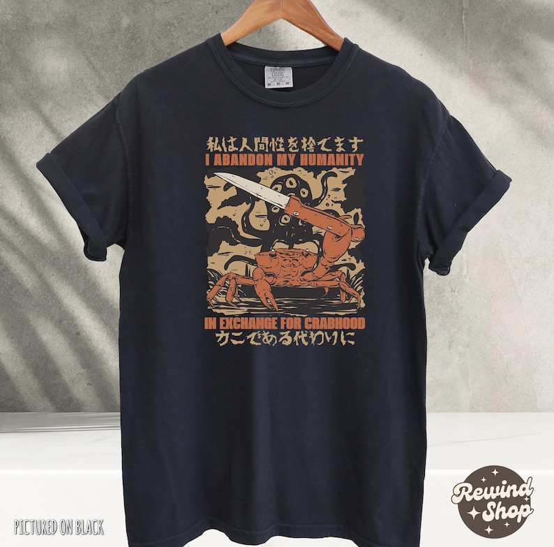 May include: Black t-shirt with a graphic featuring a crab holding a knife, with the text "I ABANDON MY HUMANITY IN EXCHANGE FOR CRABHOOD" in English and Japanese. The shirt is hanging on a wooden hanger.