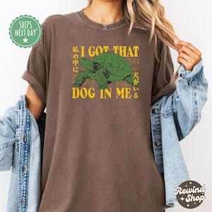 May include: Brown t-shirt with a green turtle graphic and the text "I GOT THAT DOG IN ME" in yellow. Japanese characters are also included in the design. The shirt has rolled-up sleeves.