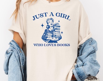 May include: A white t-shirt with blue text that reads "Just a girl who loves books". The shirt features a retro illustration of a woman holding a stack of books.