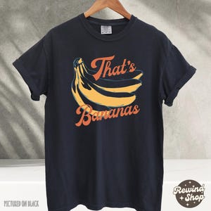 May include: Black t-shirt with a retro design featuring a bunch of yellow bananas and the text "That's Bananas" in orange. The shirt is hanging on a wooden hanger.