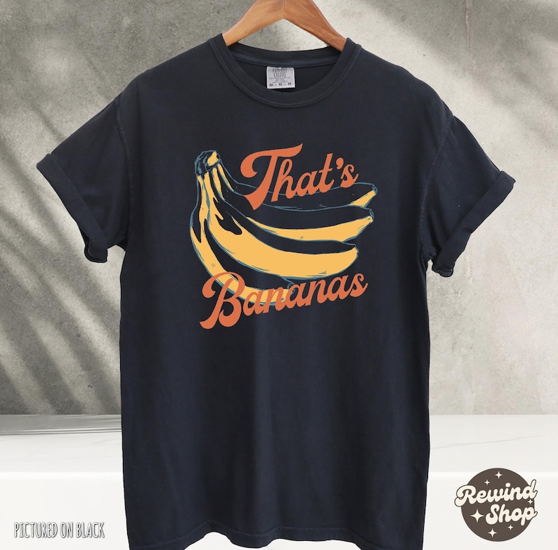 May include: Black t-shirt with a retro design featuring a bunch of yellow bananas and the text "That's Bananas" in orange. The shirt is hanging on a wooden hanger.