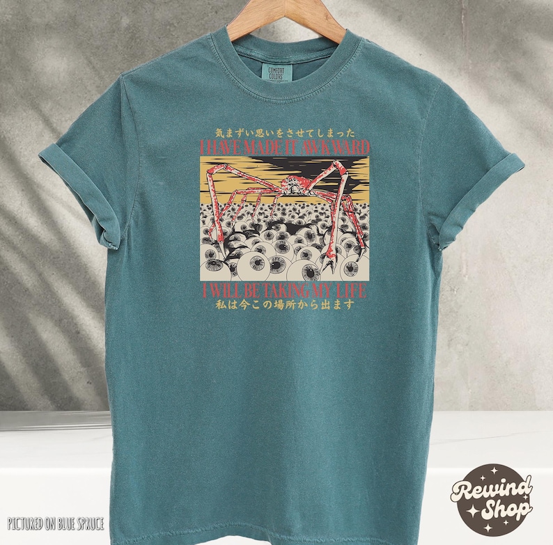 May include: Teal-colored t-shirt with a graphic illustration of a spider-like creature with red legs and a yellow body, surrounded by eyeballs. The shirt has Japanese text and the phrases "I HAVE MADE IT AWKWARD" and "I WILL BE TAKING MY LIFE".
