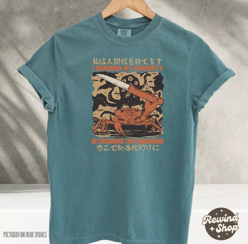May include: Teal-colored t-shirt with a graphic featuring a crab holding a knife, an octopus, and text in English and Japanese. The text reads "I abandon my humanity" and "In exchange for crabhood".