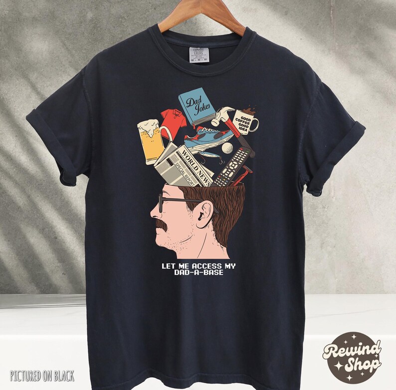 May include: Black t-shirt with a graphic of a man's head, open to reveal various items like a beer mug, sneakers, and a remote control. The text "LET ME ACCESS MY DAD-A-BASE" is below the head.