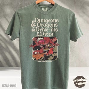 Może przedstawiać: Szałwiowa zielona koszulka z napisem "Dungeons & Dragons & Drive-ins & Dives" nad grafiką czerwonego smoka w okularach przeciwsłonecznych. Grafika zawiera r&oacute;wnież napis "Escape From Flavortown". Koszulka jest prezentowana na wieszaku.