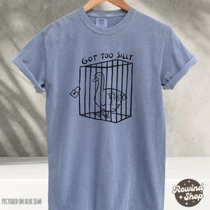 May include: A light blue t-shirt with a black graphic of a goose in a cage. The text "Got Too Silly" is printed above the graphic.