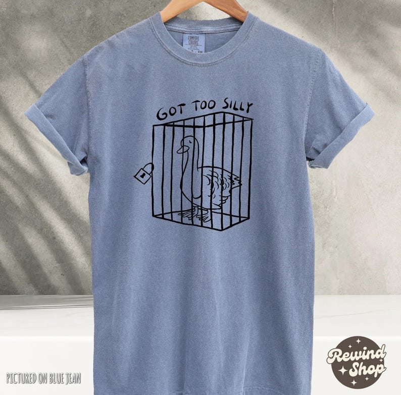 May include: A light blue t-shirt with a black graphic of a goose in a cage. The text "Got Too Silly" is printed above the graphic.